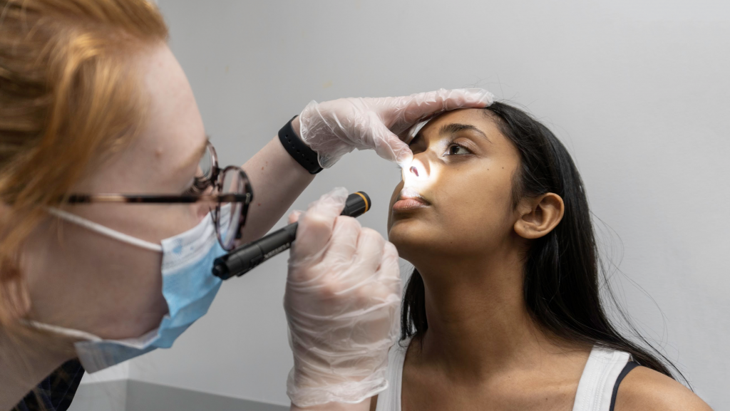 Healthcare provider has gloved thumb on client's nose and fingers on forehead to look into the nasal cavity with a penlight. Client's head is tilted slightly back.