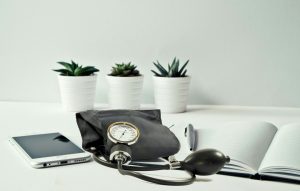 In the foreground, a black blood pressure cuff with a bulb for inflation is placed between a smartphone and a journal opened to a blank page with a pen atop it (as though ready for monitoring blood pressure levels).