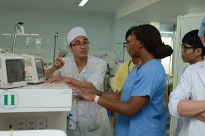 5 hospital staff including doctors and nurses gather around medical equipment in a hospital. One of the doctors is gesticulating while others listen during rounds.