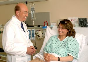 An adult woman hospital patient is sitting up in her hospital bed with her hands folded on her lap. The doctor is an older man with a white lab coat; he stands with hands folded in front of him. Both the patient and doctor are smiling and the mood of the hospital room is warm and hopeful.