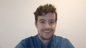 Christian Haggar, our case study patient. He has dark brown curly hair, cropped shorter at the sides than at the top. He has light facial hair and is smiling with his teeth in a blue denim button up against a white background.