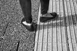Black and white photo of a person's feet with flat shoes stepping on two different textures of pavement.