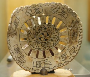 A carved marine shell featuring a spider motif at center with circle of hands with shell bracelets.