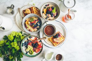 Photograph of a breakfast table. It includes waffles, fruits, cereals, milk and others.