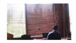 A person sitting at a desk wearing headphones while using a laptop and facing a large window on a sunny day.