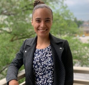 An image of Savannah Black, a Registered Dietitian and educator, stands confidently on a balcony with greenery in the background. Her open smile and poised stance reflect her passion for mentorship and inclusive, community-based health care.