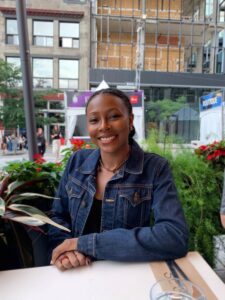 This is an image of Sierra Wellington, a young Black woman smiling as she sits at a table in front of a small garden in a city setting.