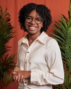 This is an image of Joie Shaw, a dedicated dietitian and researcher, stands in front of a warm-toned backdrop framed by green palms, radiating a thoughtful and welcoming presence.