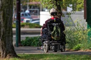 Photo of a person using an electrical wheelchair on a cement pathway lined with grass, trees, and flowers.