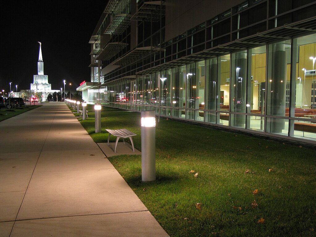 A nighttime view of Brampton Civic Hospital, with a long, well-lit sidewalk lined by modern light posts and benches. The hospital’s glass facade reflects the interior lights. A lit-up white temple-like building is visible in the background.