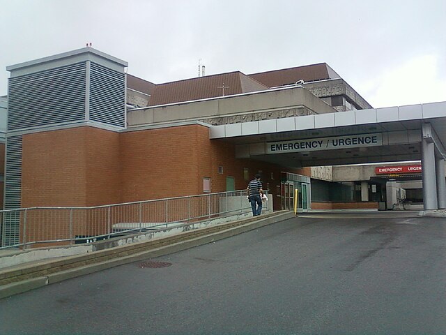 A person walks up a ramp toward the emergency entrance of the Children's Hospital of Eastern Ontario in Ottawa. The entrance sign reads “EMERGENCY / URGENCE,” and the building is made of brick and concrete.
