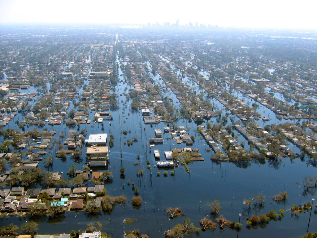 Aerial view of a flooded residential neighborhood in New Orleans after Hurricane Katrina, with streets submerged and houses surrounded by water. The city skyline is faintly visible in the background.