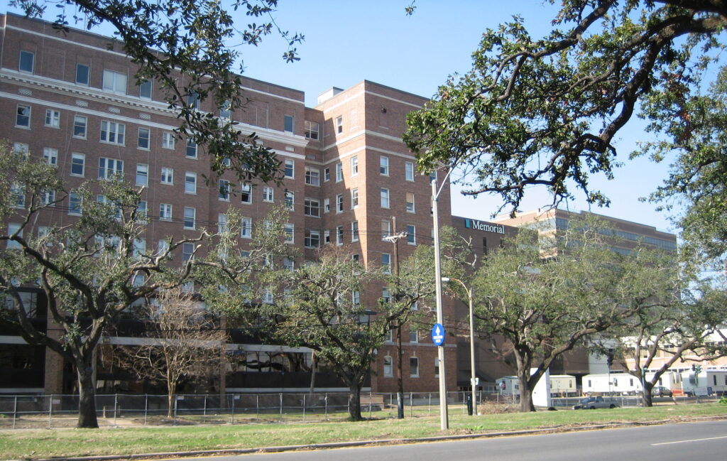 Exterior of Memorial Medical Center in New Orleans after Hurricane Katrina, showing a large brick hospital building with broken windows and several temporary trailers parked outside. Leafless trees and debris are visible in the foreground.