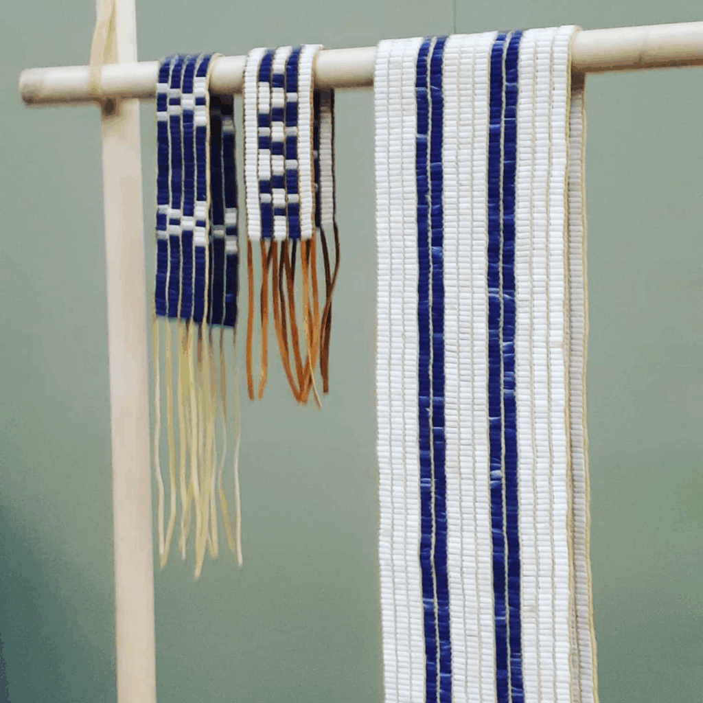 Four wampum belts hang from a wooden rod. The belts are made of white and dark blue beads arranged in traditional patterns, each representing different treaty agreements. The largest belt has two thick dark blue vertical stripes, while the others feature geometric designs. The belts are displayed against a pale green background.