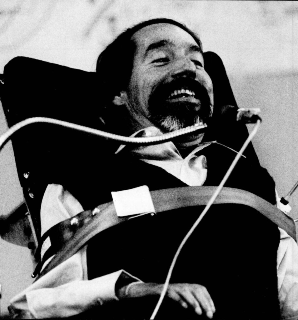 Black and white photo of Ed Roberts, a disabled man with a beard and mustache, smiling while seated in a power wheelchair. He is wearing a dark vest over a light shirt, and a seatbelt crosses his chest. A breath control tube is visible near his face, used to operate the wheelchair.