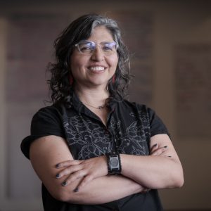 A Photograph of Andrea Parra, a woman with tan skin, brown and grey hair. She is standing with her arms crossed, smiling at the camera. She is wearing clear-framed glasses, a digital watch, a black shirt, and blue nail polish.