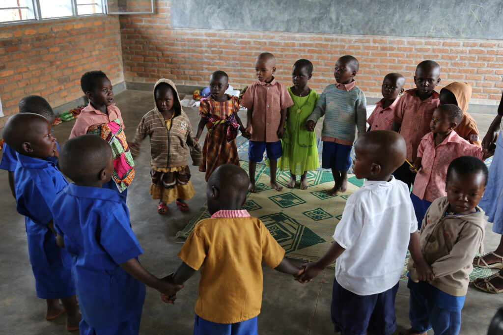 A group of children holding hands and singing in a rural Rwandan preschool classroom.