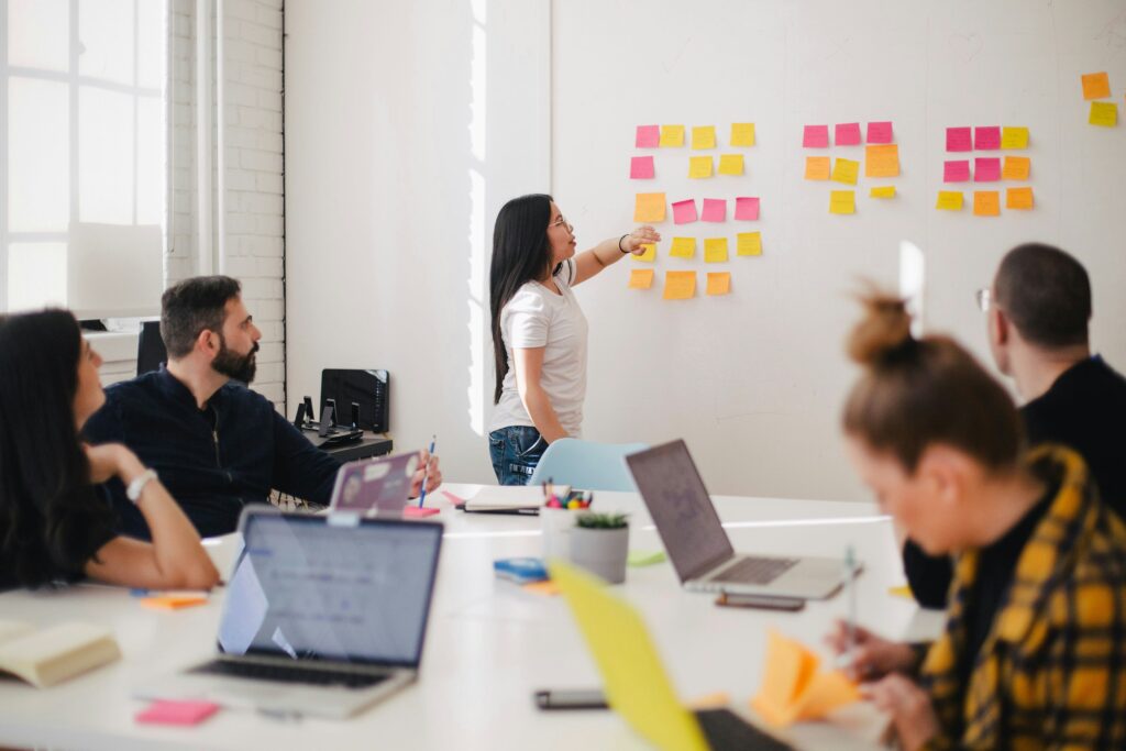 group at a desk with laptops with one person standing at whiteboard putting up sticky notes