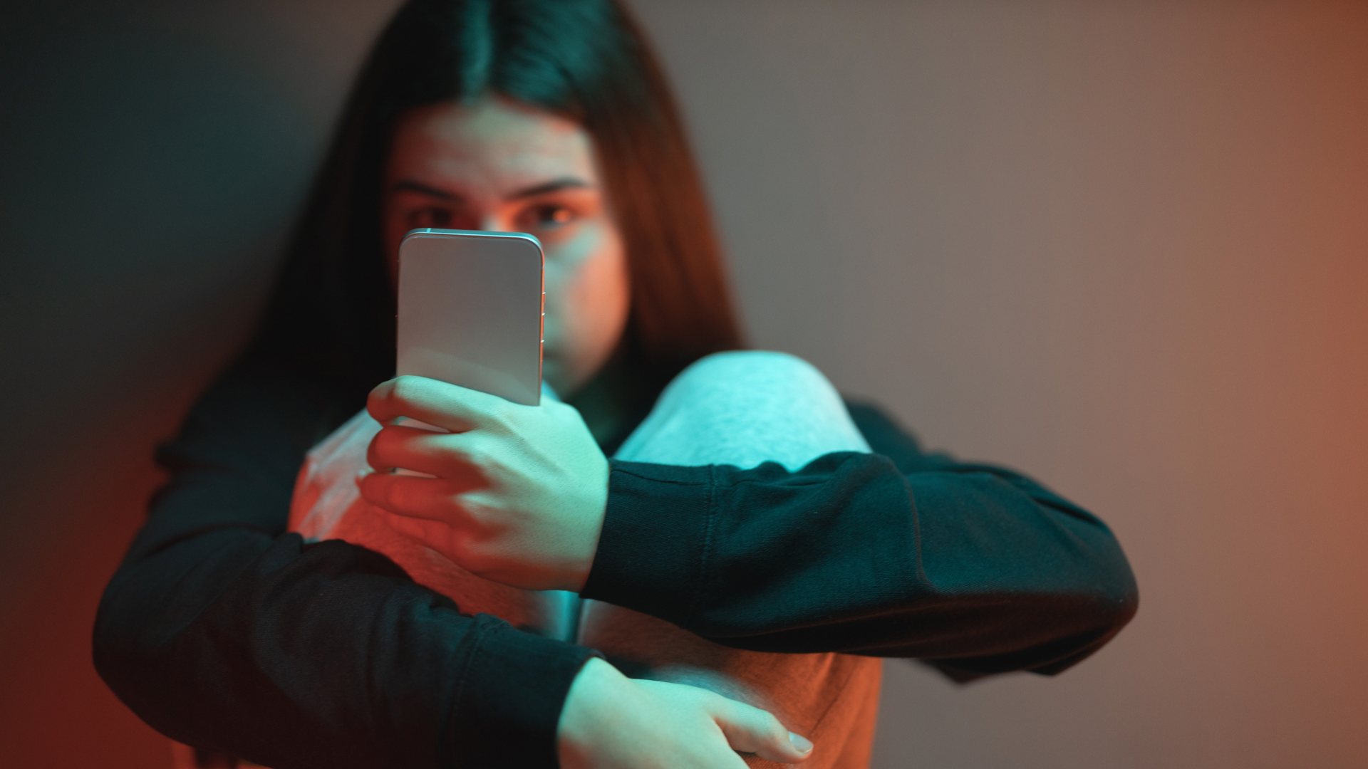 A person sits with their knees drawn up, holding a smartphone close enough to hide their face.